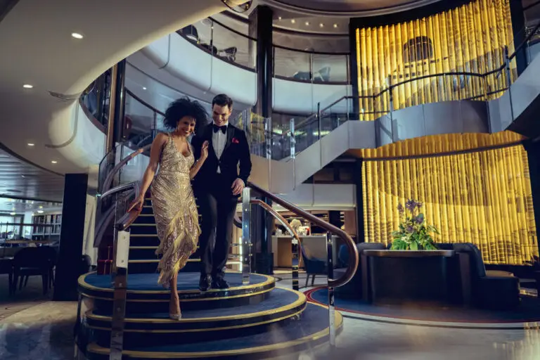 Couple in formal attire walking down the main stairs in Cunard Queen Anne's grand lobby on gala night