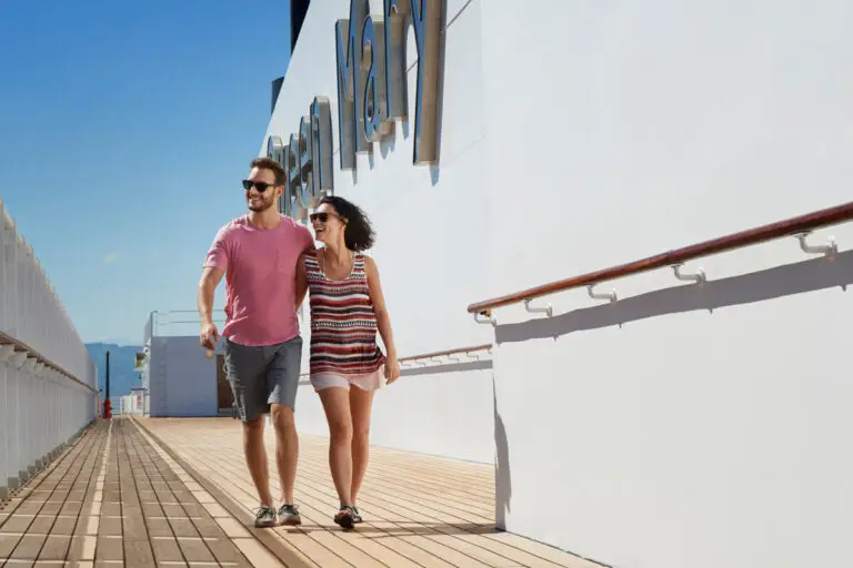 A man and a woman walk arm in arm on the deck of Cunard's Queen Mary 2, smiling and enjoying a sunny day. The ship’s white exterior and railings add to the elegance of this luxury cruise under the clear blue sky.