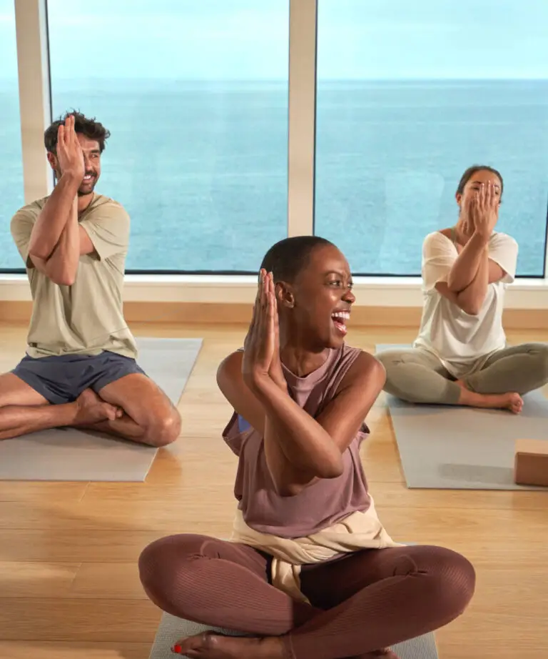 Three people sit on yoga mats indoors, performing a seated yoga pose with arms intertwined, as large windows reveal a sea view, experiencing B-Complex Balance onboard Virgin Voyages. All appear relaxed and focused.