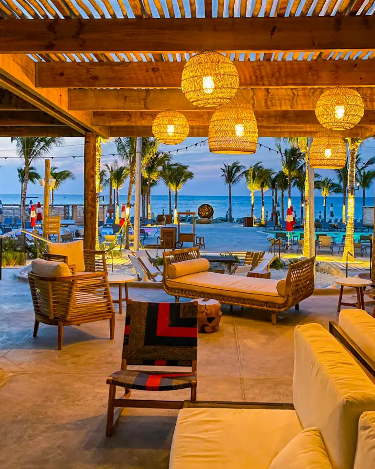 A cosy outdoor lounge with wooden furniture and woven lamps at Virgin Voyages The Beach Club overlooks palm trees and a beach at sunset, creating a warm, inviting atmosphere.