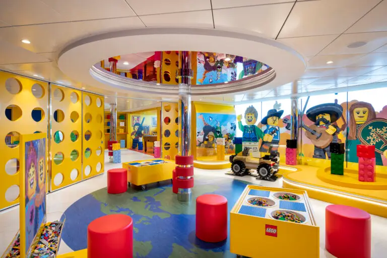 A colourful indoor playroom on MSC Virtuosa with yellow walls, LEGO themed decorations, large LEGO figures, and a fun mini club vibe. Complete with activity tables, red stools, and playful art designed for children’s creativity and activities.