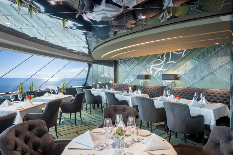 Elegant MSC Cruises Yacht Club restaurant on MSC Virtuosa with large windows overlooking the sea, round tables with white tablecloths, grey upholstered chairs, and modern wall art. Each table is set with glassware, serviettes, and small flower arrangements.