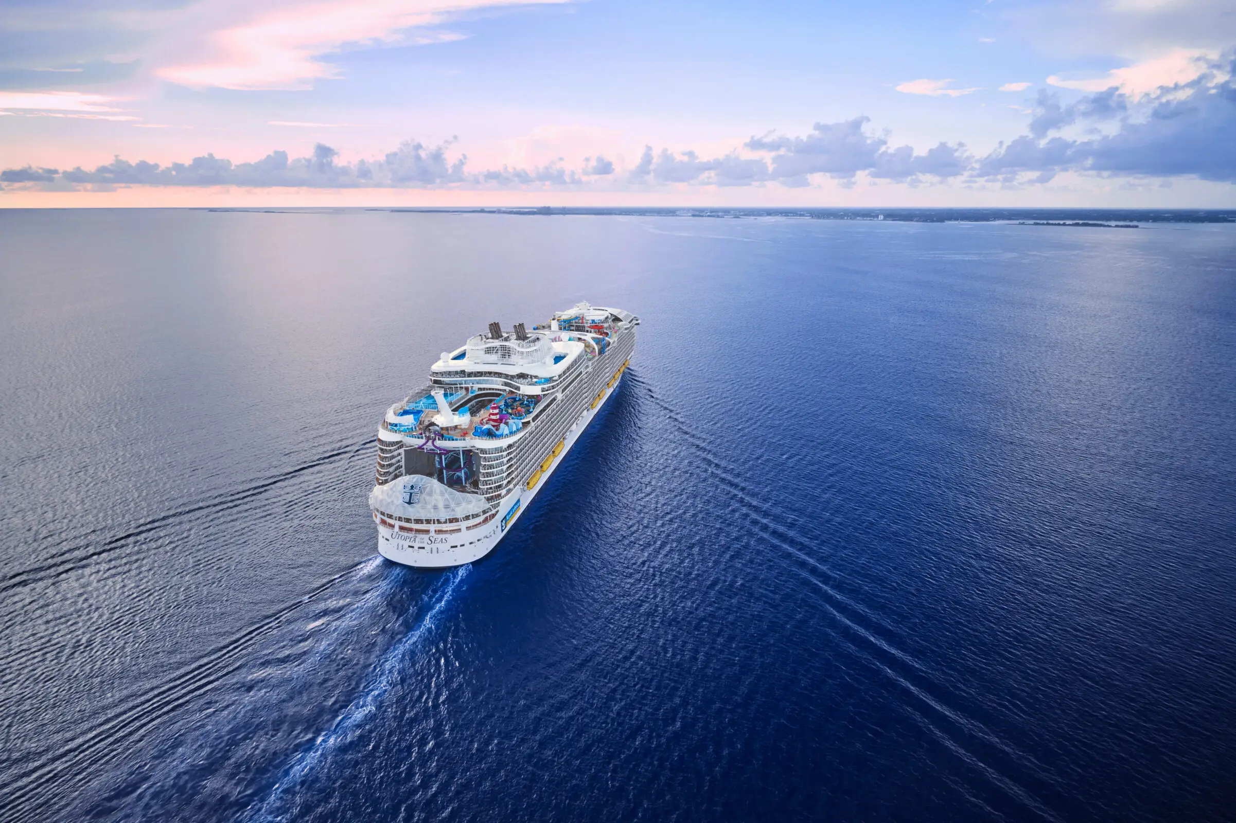 Royal Caribbean's Utopia of the Seas sails across calm, deep blue ocean waters under a pastel-coloured sky at sunset, leaving a gentle wake behind it.