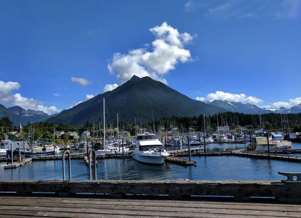 A marina with boats docked, set against a backdrop of a large, green mountain under a blue sky with scattered clouds.