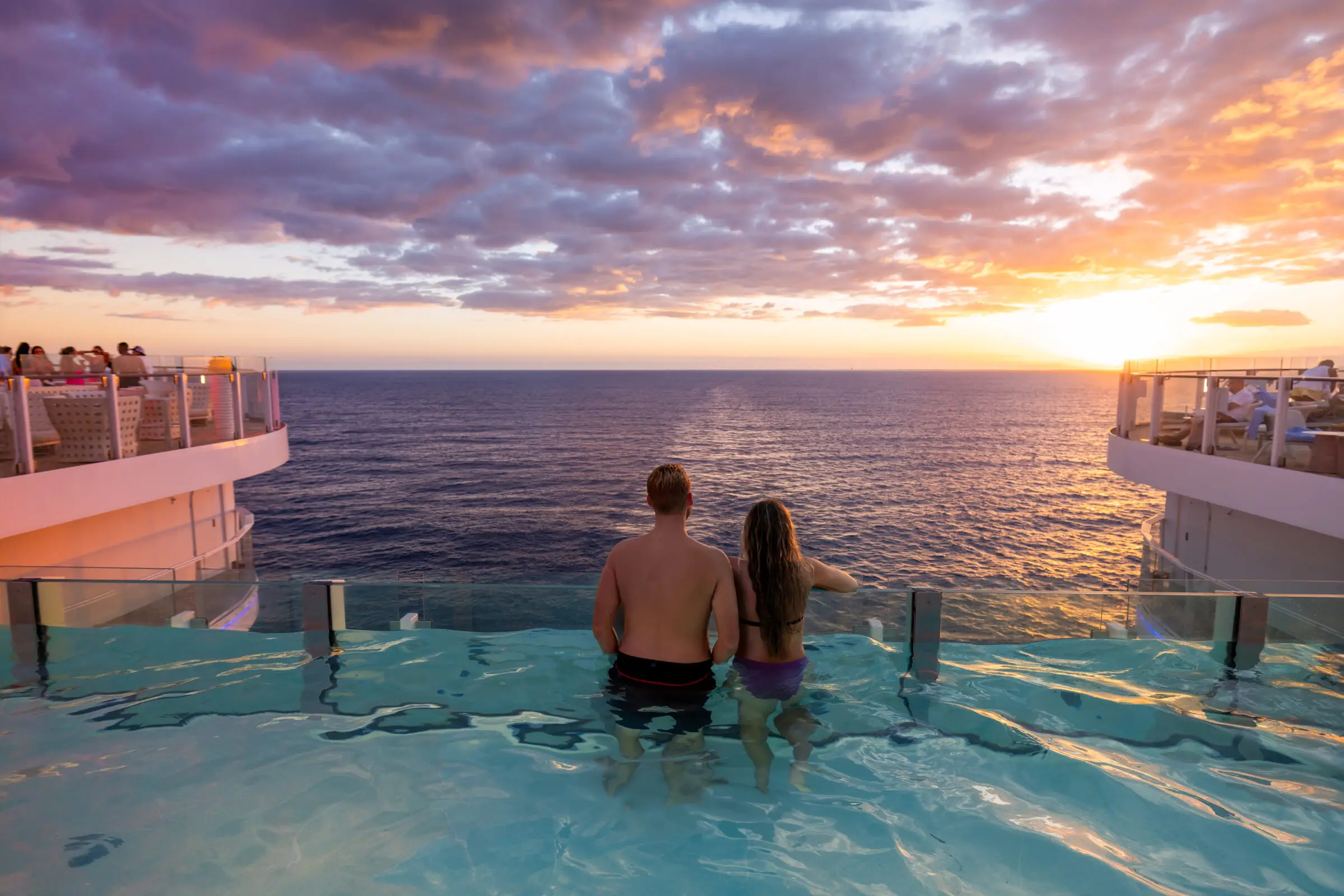 A couple stand together in The Hideaway Pool on a Royal Caribbean cruise ship, facing the sea from the infinite pool and watching a colourful sunset under a partly cloudy sky. Other passengers are visible on deck in the background.