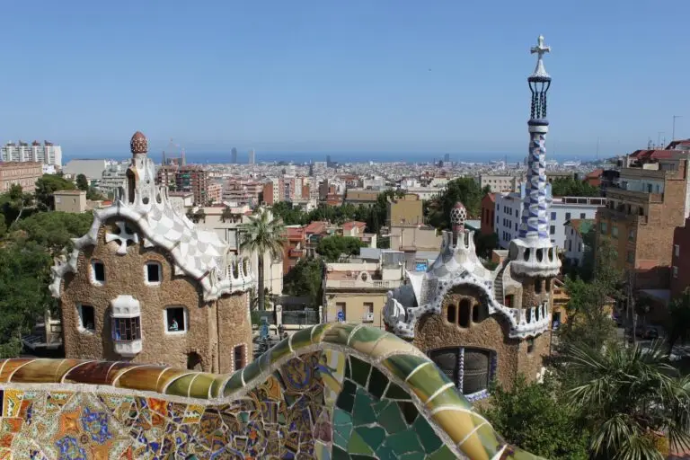A panoramic view of Barcelona from Park Güell shows colourful mosaic tiles in the foreground and whimsical Gaudí-designed buildings, with the vibrant city and sea stretching out in the background under a clear blue sky.