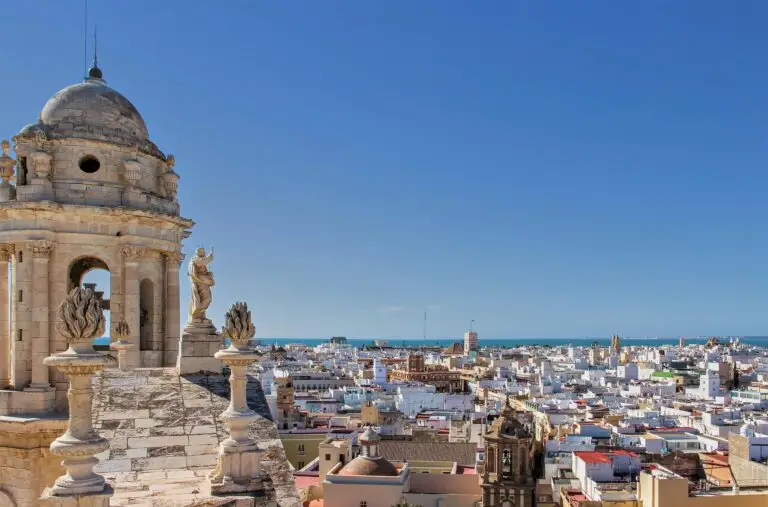 A view from a historic stone cathedral with statues in Cádiz, overlooking a city of white buildings and rooftops, with the blue sea and clear sky in the background.
