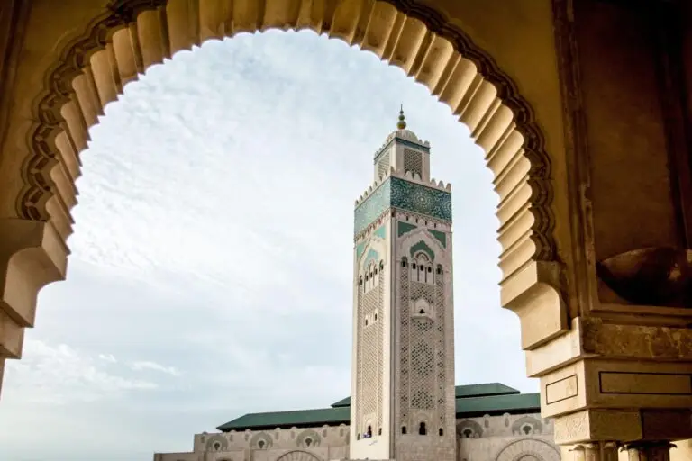 The image shows a tall, ornate mosque minaret in Casablanca, framed by an archway with intricate carvings, set against a partly cloudy sky.