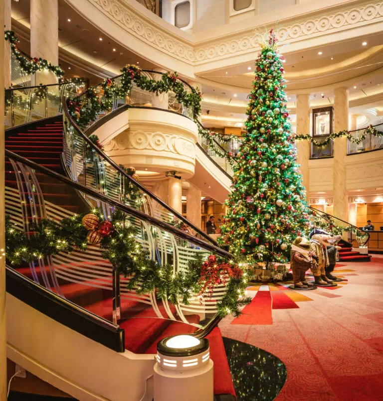 A grand Cunard lobby decorated for Christmas, featuring a large, decorated Christmas tree, festive garlands on a sweeping staircase, red carpet, and warm lighting creating a luxurious, holiday atmosphere.