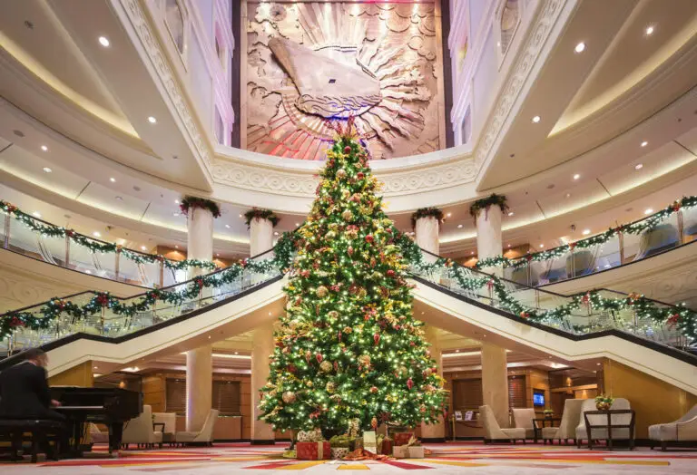 A large, decorated Christmas tree stands in the centre of Cunard’s grand, elegant lobby with sweeping staircases, festive garlands on railings, gifts under the tree, and a large artwork on the wall above.