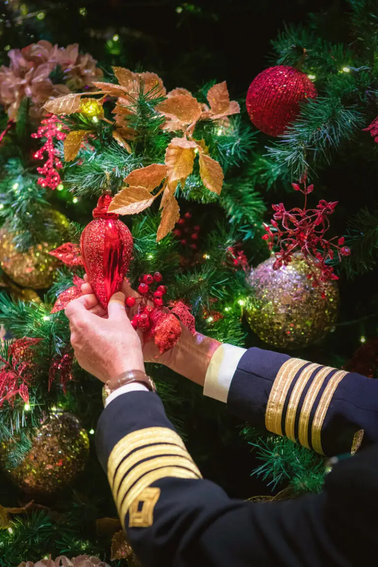 A officer in a dark Cunard uniform with gold stripes on the sleeves decorates a Christmas tree with red and gold baubles and glittering leaves.