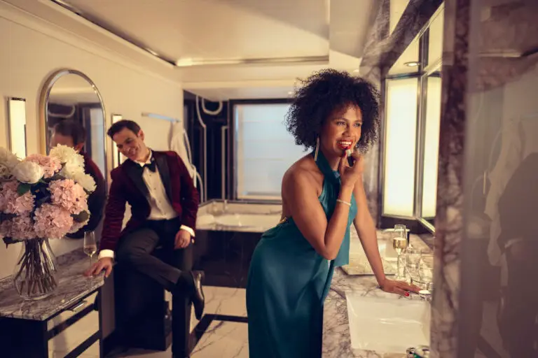 A woman in a teal dress applies lipstick in a luxurious Cunard grills suite bathroom, smiling at her reflection. A man in a dinner jacket leans against the counter in the background, watching her with a relaxed expression.