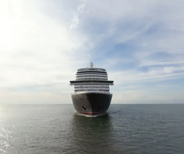 The Cunard Queen Anne, a large cruise ship, sails directly towards the camera on calm, open water under a partly cloudy sky.