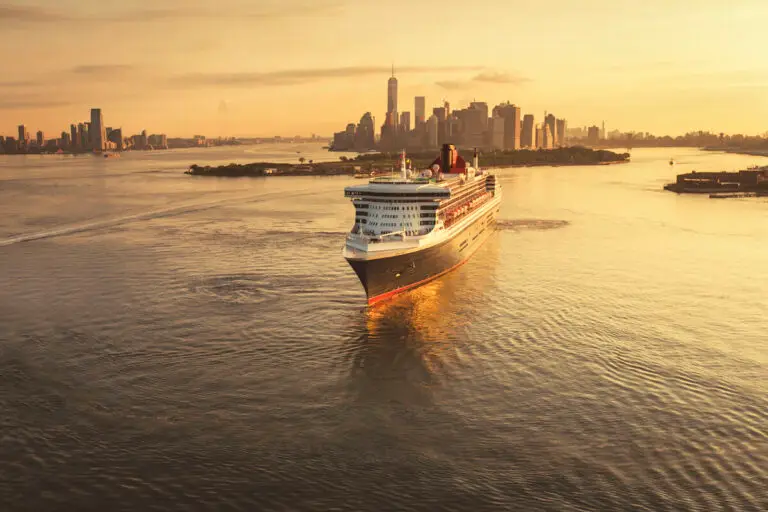 The Cunard Queen Mary 2 sails away from the New York City skyline at sunset, with warm golden light reflecting on the water and tall buildings visible in the distance.