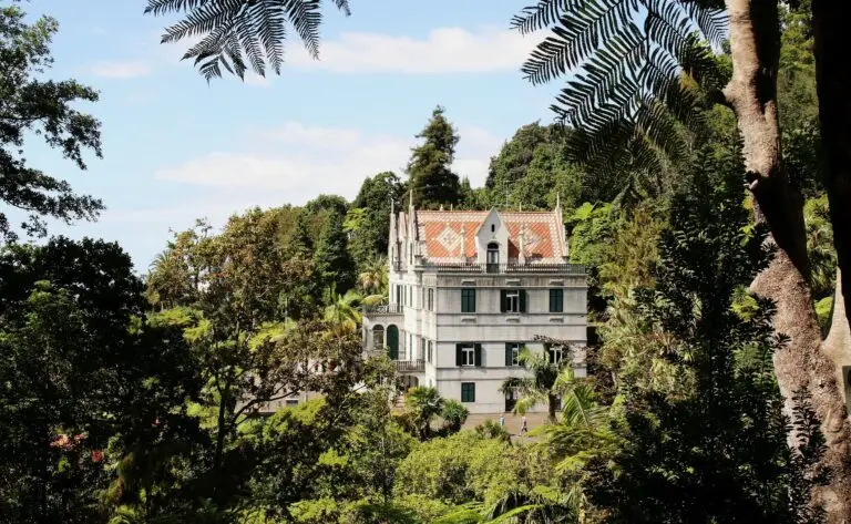 A large white mansion with orange roof tiles stands surrounded by lush green trees and foliage under a blue sky with scattered clouds, evoking the elegant charm of Funchal.