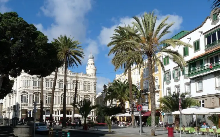 A sunny square in Gran Canaria with palm trees, outdoor café tables, and historic buildings with white façades and balconies under a bright blue sky with scattered clouds.