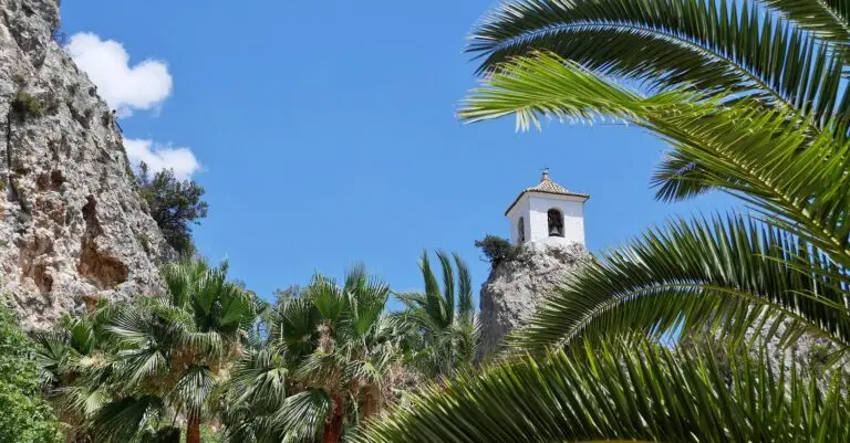A small white bell tower stands atop a rocky cliff in Guadalest, Alicante, Spain, surrounded by green palm trees, against a bright blue sky with a few scattered white clouds.
