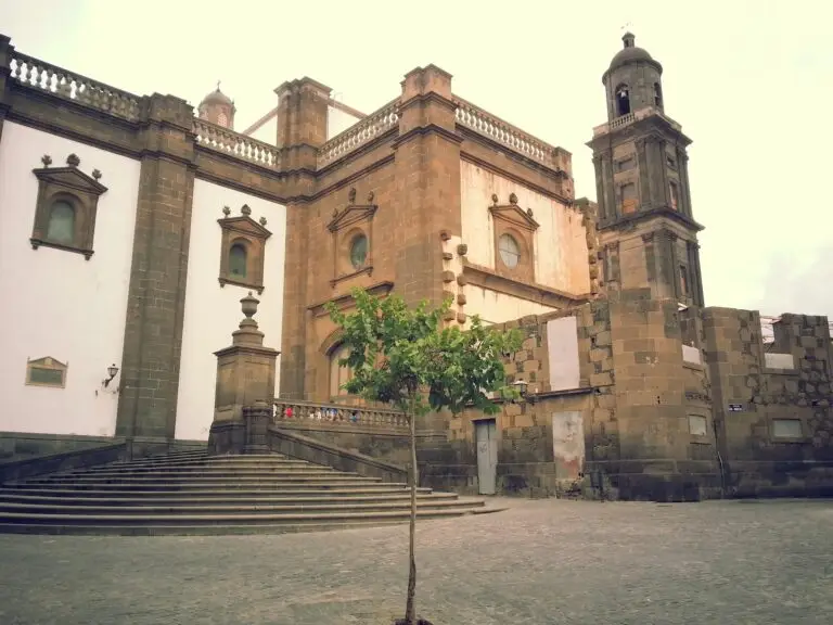 Historic stone building with arched windows and a bell tower, set beside a wide staircase and a small tree in the foreground, located in a cobbled plaza of Las Palmas de Gran Canaria under a cloudy sky.