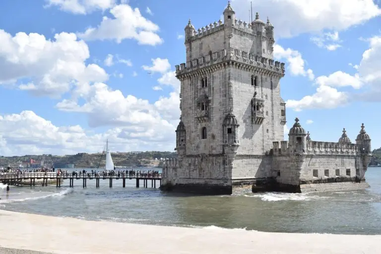 A historic stone tower, the Lisbon Belém Tower, stands by the water’s edge with a bridge connecting it to the shore. People walk along the bridge, and a white sailing boat is visible in the background under a partly cloudy sky.