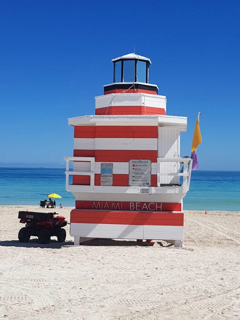 A red and white striped lifeguard tower with a black roof stands on Miami Beach’s sandy shore. A small vehicle is parked beside it and a yellow umbrella with people can be seen in the background, with the blue Miami sea and sky in the distance.