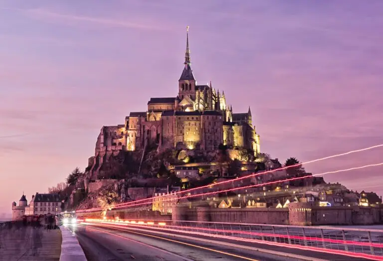 Mont Saint-Michel, a historic island abbey in France, is illuminated at dusk with a purple sky. Light trails from vehicles are visible on the road leading to the abbey.