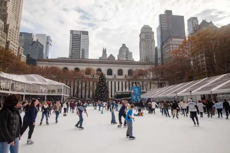 People are ice skating at an outdoor rink in New York, surrounded by tall buildings and autumn trees. A large decorated Christmas tree stands in the centre, with a historic building visible in the background under a cloudy sky.