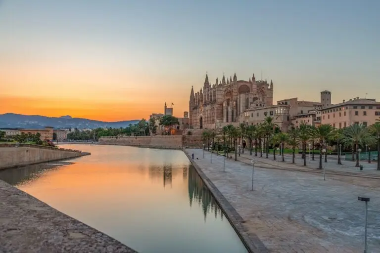 A view of the large Gothic cathedral in Palma de Mallorca near a reflecting pool at sunset, with palm trees lining the promenade and mountains visible in the distance under an orange sky.