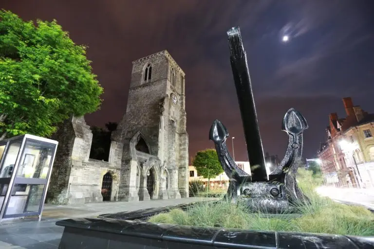 The large black QE2 anchor stands in front of the Holyrood historic stone church ruin in Southampton at night, with a tall clock tower, arched windows, and nearby buildings under a cloudy, moonlit sky.