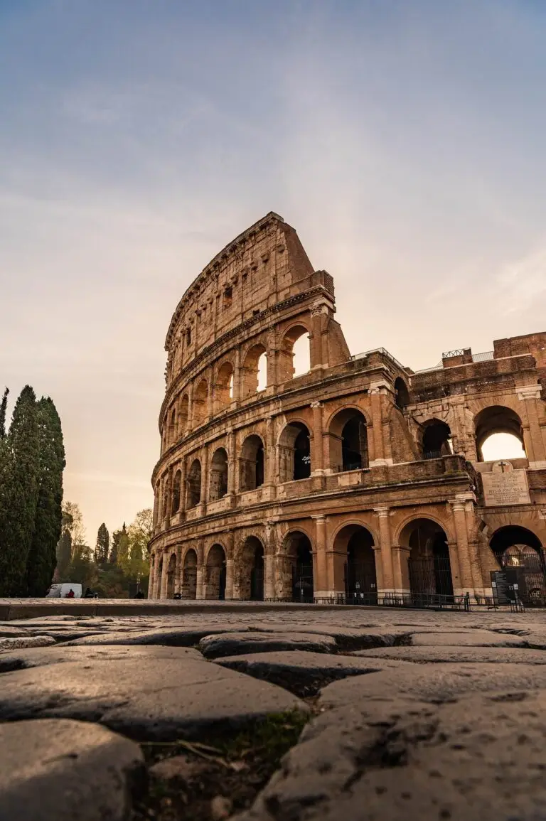 The image shows the Colosseum in Rome, Italy, captured from a low angle at sunrise or sunset, with stone pavement in the foreground and a clear sky in the background.