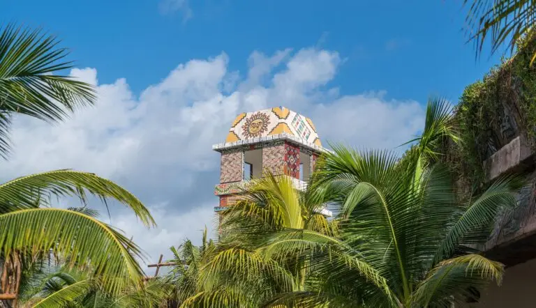 A colourful, patterned tower with a decorative sun motif rises above lush green palm trees in Costa Maya, set against a bright blue sky with scattered clouds.