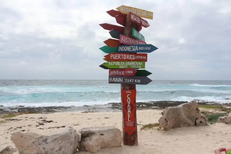 A colourful signpost on a sandy beach in Cozumel, Mexico, points to various global destinations with distances. Ocean waves lap the shore under a cloudy sky, with rocky formations adding charm to this scenic Cozumel coastline.