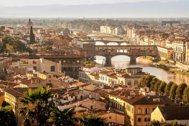 A panoramic view of Florence, Italy, featuring the Ponte Vecchio bridge spanning the Arno River, surrounded by historic buildings with terracotta roofs, and distant rolling hills under a hazy sky.