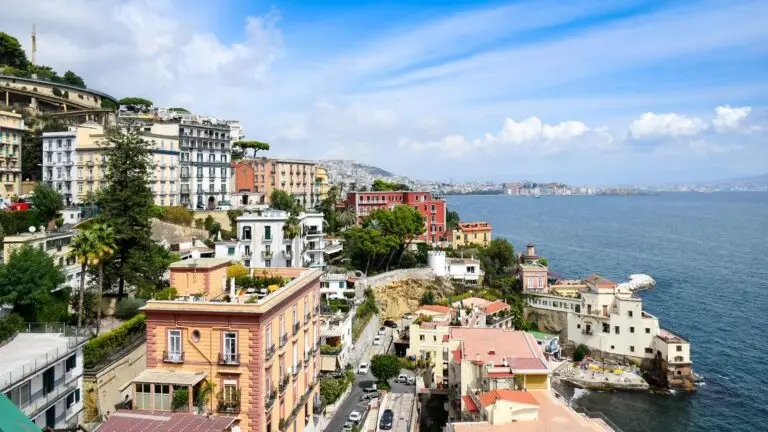 Colourful buildings line the Naples coastal hillside overlooking the blue sea, with winding roads and lush greenery, under a bright sky with scattered clouds. The background shows distant cityscape and mountains across the water.