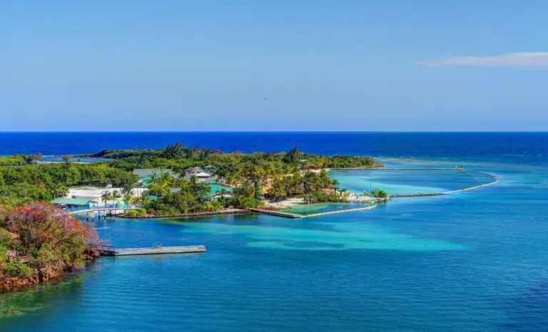Aerial view of Roatán with lush greenery, scattered buildings, and jetties, surrounded by clear turquoise and deep blue waters under a bright blue sky.