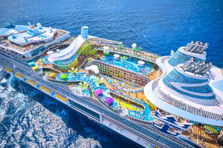 Aerial view of the Royal Caribbean Icon of the Seas, a colourful cruise ship sailing on the ocean, featuring multiple pools, water slides, sun decks, and recreational areas filled with people enjoying amenities under clear blue skies.