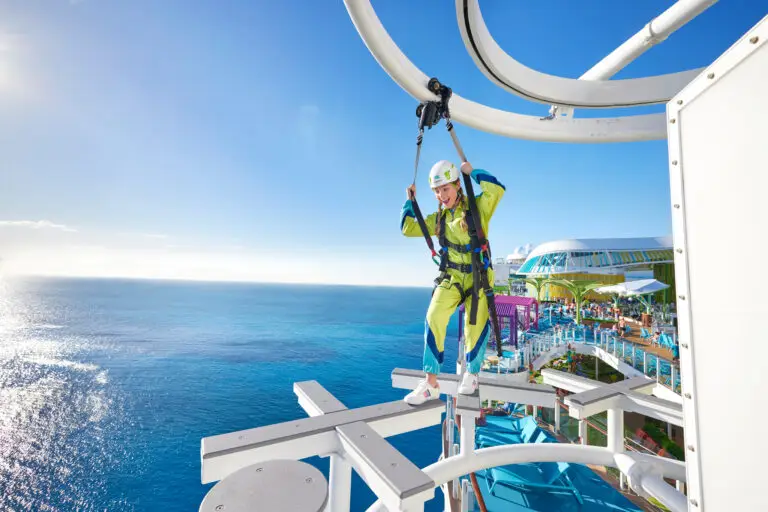 A person wearing a harness and helmet walks on a narrow plank high above the Royal Caribbean cruise ship deck, experiencing the thrilling Crown’s Edge with a bright blue ocean and clear sky in the background.