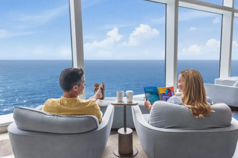 A man and woman sit in chairs at the Pearl Cafe, looking out a window at the ocean aboard their Royal Caribbean cruise.